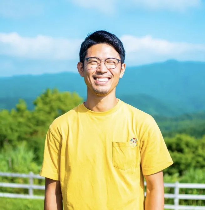 Mr. Akai smiling outdoors, with green mountains behind him
